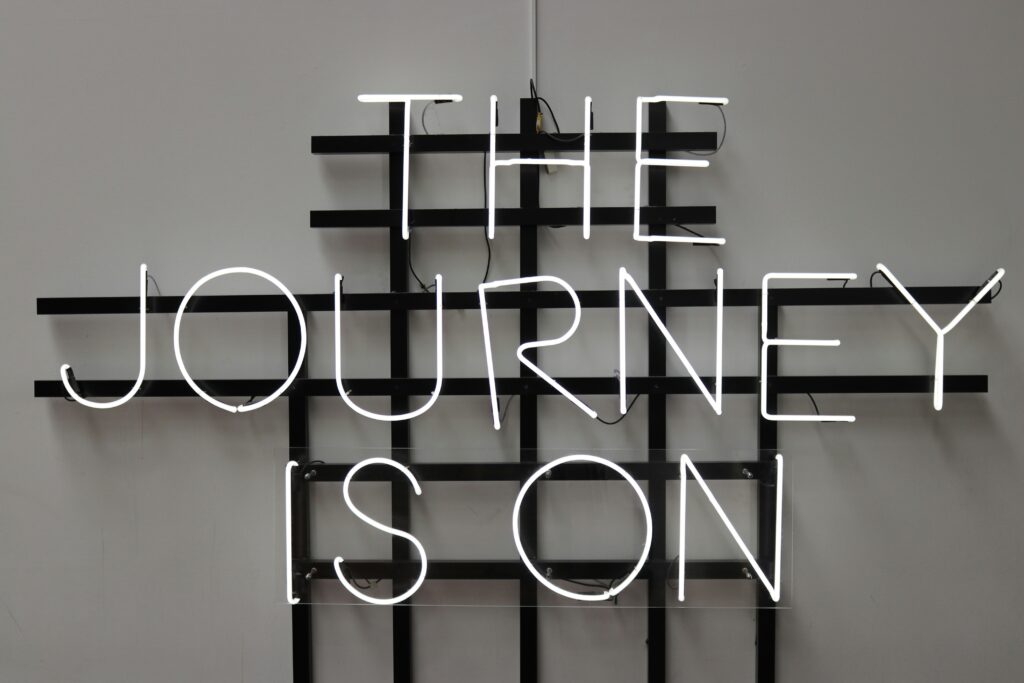 Neon sign with white light reading "THE JOURNEY IS ON" against a black grid background on a neutral wall, conveying a motivational and adventurous tone.
