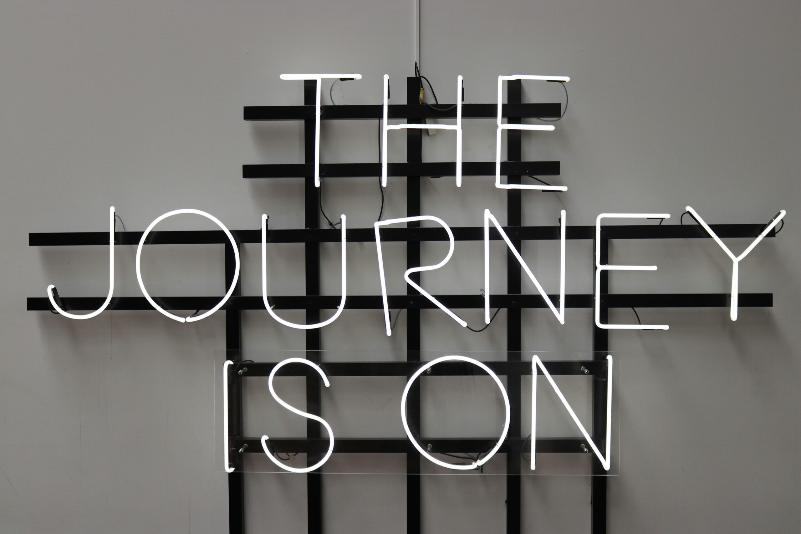 Neon sign with white light reading "THE JOURNEY IS ON" against a black grid background on a neutral wall, conveying a motivational and adventurous tone.