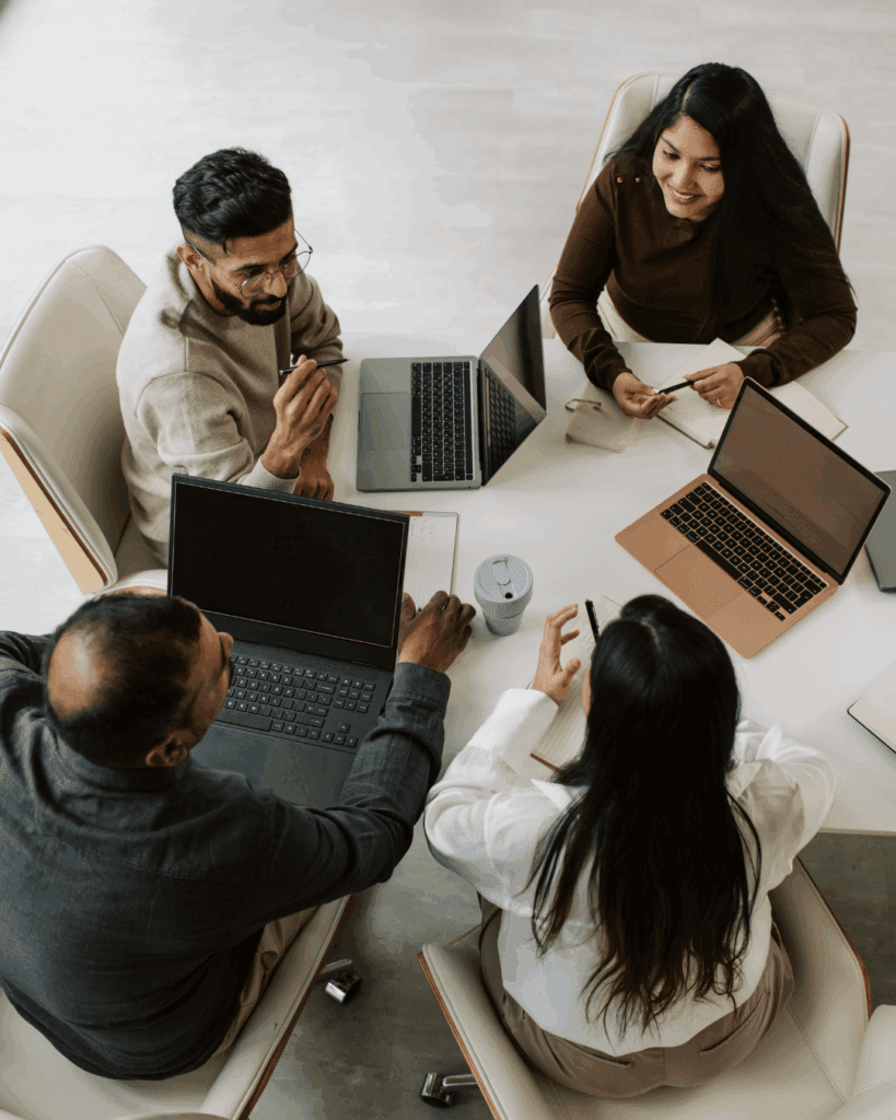 Four professionals engage in a collaborative meeting, discussing ideas around laptops on a conference table. A coffee cup is visible, highlighting a casual work environment.