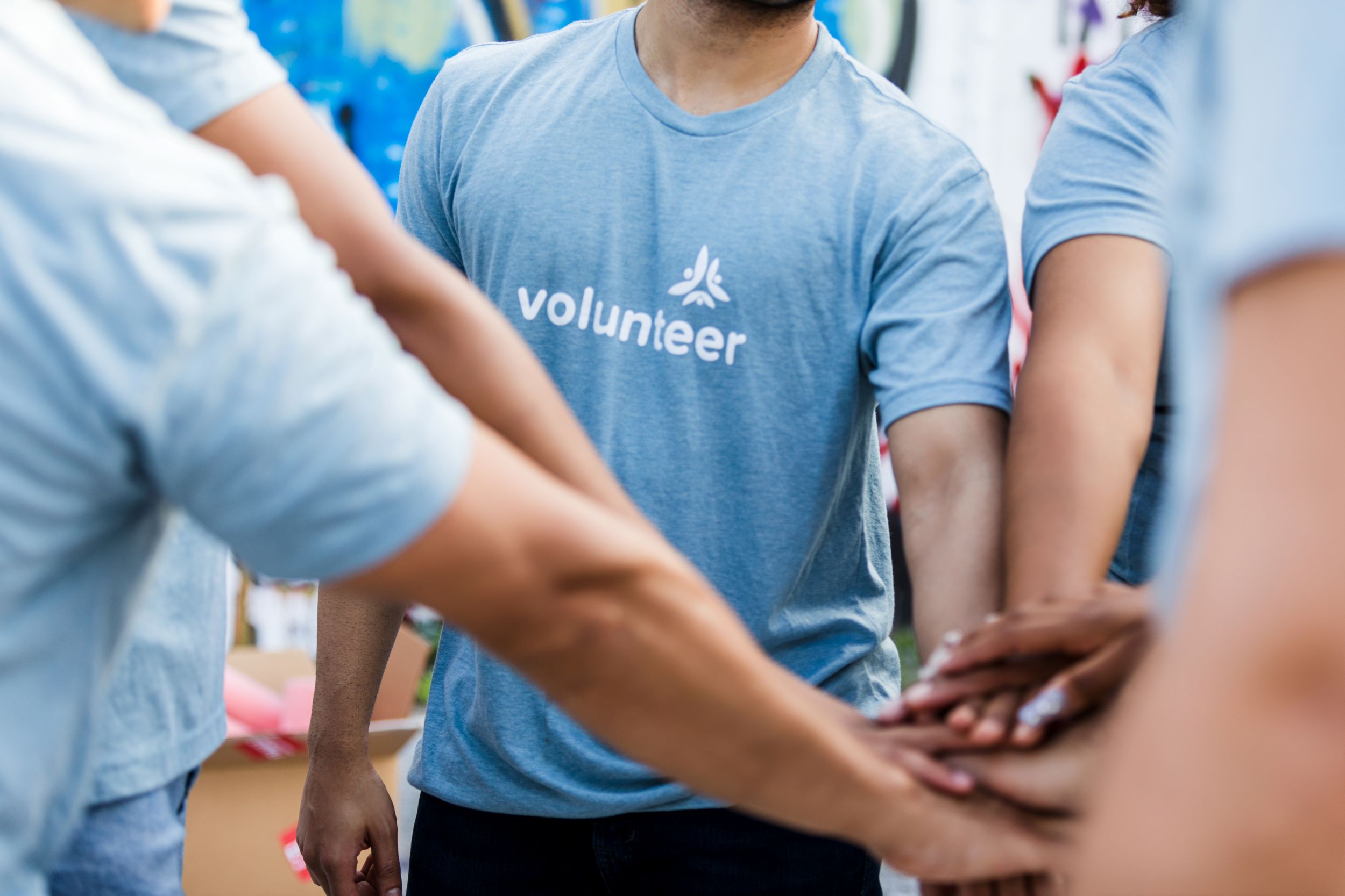 A group of volunteers in matching blue shirts share a unified moment by stacking their hands together, symbolizing teamwork and community engagement.