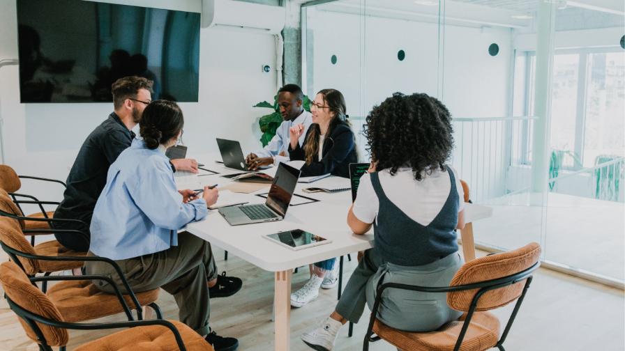 A diverse team collaborates around a conference table, discussing strategies and sharing ideas, with laptops and notepads in a modern office setting.