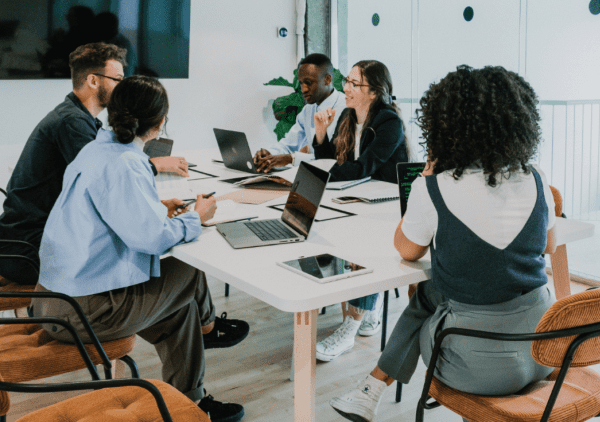 A diverse group of six professionals engages in a collaborative meeting at a modern conference table, laptops open and ideas being shared.